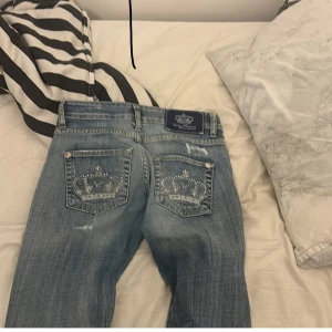 Blå jeans med broderade detaljer - Säljer ett par snygga blå jeans med coola broderade kronor på bakfickorna. De har en klassisk femficksdesign och en lätt slitning framtill för en trendig look. Perfekta för vardagsbruk eller en avslappnad utekväll!