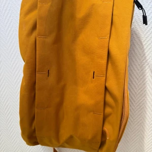 Ryggsäck - Aldrig använd! Part backpack, part messenger bag, inspired by base rigs. The Hugger Base is the pack that meets you halfway - literally. In backpack mode, one tug of the D-ring deploys the bag into an on-body, side-access messenger pack. 15L