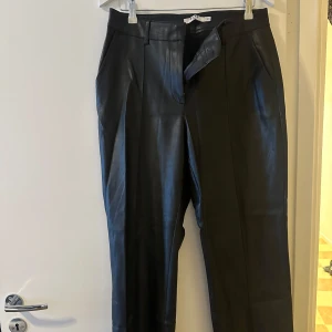 Svarta byxor från NA-KD - Säljer ett par snygga svarta läder byxor från NA-KD. De är högmidjade och har en stilren design med knappar och dragkedja. Perfekta för både vardag och fest!