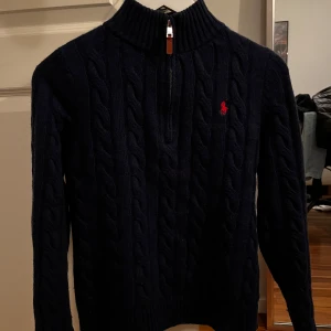 Mörkblå stickad tröja från Polo Ralph Lauren - Säljer en snygg mörkblå stickad tröja från Polo Ralph Lauren med kabelstickat mönster och en halv dragkedja framtill. Tröjan har en broderad röd logotyp på bröstet och långa ärmar. Perfekt för kyliga dagar!
