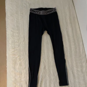 Svarta träningsleggings från Craft - Säljer ett par svarta träningsleggings från Craft med grå detaljer och elastisk midja. Perfekta för löpning eller gymmet. Bekväma och stretchiga för optimal rörelsefrihet.