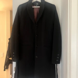 Svart kappa från H&M - Elegant svart kappa från H&M med knappar framtill och klassisk krage. Den har långa ärmar och ett bälte i midjan för en stilren look. Perfekt för kyligare dagar. Från H&M.