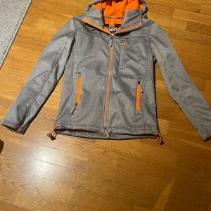 Grå vindjacka från Superdry - Säljer en grå vindjacka från Superdry med orange detaljer och huva. Jackan har dragkedjor och är perfekt för blåsiga dagar. Den har en sportig look med långärmade ärmar och justerbara snören vid nederkanten.