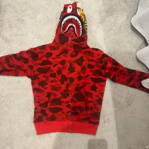 Röd camo hoodie med hajmotiv - Storlek XL men passar som M💗Säljer en unik röd camo hoodie med ett coolt hajmotiv på huvan. Hoodien har dragkedja och är perfekt för att sticka ut i mängden. Den har långa ärmar och en bekväm passform.
