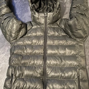 Svart dunjacka från Peak Performance - Snygg grön dunjacka från Peak Performance med huva och dragkedja. Perfekt för kyliga dagar med sin quiltade design som håller värmen. Jackan har en stilren look och passar bra till vardags.