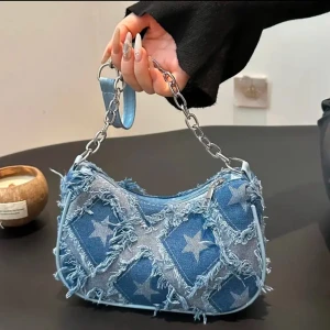 Blå jeansväska med stjärnor - Cool axelväska i blått jeansmaterial med fransiga detaljer och stjärnmönster. Väskan har en kedjerem och en dragkedja för säker förvaring. Perfekt för en trendig look!