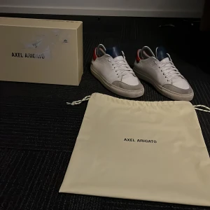 Axel Arigato Clean 90 triple - Snygga vita sneakers från Axel Arigato med röda och blå detaljer. Skorna har en klassisk design med snörning och kommer med originalförpackning och dustbag.