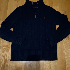 Mörkblå stickad tröja från Polo Ralph Lauren - Säljer en snygg mörkblå stickad tröja från Polo Ralph Lauren med kabelstickat mönster och en röd broderad logotyp på bröstet. Tröjan har en dragkedja vid halsen och ribbade muddar.