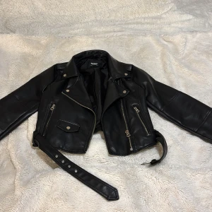Svart ”skinnjacka” - Snygg svart skinnjacka från Pull&Bear med dragkedjor och bälte. Perfekt för en cool look med sina detaljerade fickor och klassiska design. Passar utmärkt till både vardag och speciella tillfällen. Få använda gånger. Stl 38