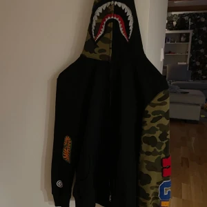Bape tröja - Säljer en unik hoodie med hajmotiv på huvan och camouflageärmar. Den har färgglada detaljer och en cool design som sticker ut. Perfekt för dig som vill ha något annorlunda i garderoben! Passar även xs