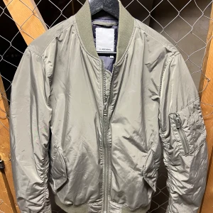 Olivgrön bomberjacka från J.Lindeberg - Snygg grå bomberjacka från J.Lindeberg med dragkedja och ribbade muddar. Jackan har en klassisk design med fickor och en ärmficka med dragkedja. Perfekt för en stilren look. Använd vid ett par få tillfällen