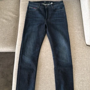 Mörkblå jeans från Tommy Jeans - Snygga mörkblå jeans från Tommy Jeans med klassisk femficksdesign och diskret logga på bakfickan. Perfekta för en stilren look. Midwaist och straight passform.