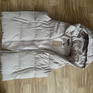 Beige dunväst från Shein - Säljer en ny och snygg beige dunväst från Shein med dragkedja och huva. Västen har en enkel design med två fickor framtill.