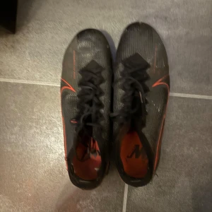 Svarta fotbollsskor från Nike - Säljer ett par svarta fotbollsskor från Nike med röda detaljer och snörning. Lite sönder sletna på enstaka ställen men går perfekt att spela i dem