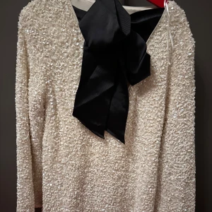 Vit glittrig klänning med stor svart rosett - Elegant vit klänning täckt av glittriga paljetter. Den har långa ärmar och en stor svart rosett framtill som ger en lyxig touch. Perfekt för festliga tillfällen.