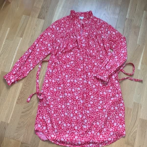 Röd blommig klänning från Holly & Whyte - Charmig röd klänning med blommigt mönster i recycled polyester från Holly & Whyte. Den har långa ärmar och ett knytband i midjan för en smickrande passform. Perfekt för en färgglad look!