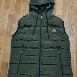 Moncler-väst - Snygg grön dunväst från Moncler med huva och dragkedja. Västen har Moncler-logga på bröstet och text längs insidan av huvan.