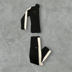 Svart set från Nike - Snyggt svart träningsset från Nike med vita detaljer. Jackan har dragkedja och långa ärmar, medan byxorna har elastisk midja och vita ränder längs sidorna. Perfekt för sportiga aktiviteter.