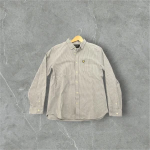 Grå långärmad skjorta med broderad detalj - Snygg grå långärmad skjorta med en broderad detalj på bröstfickan. Skjortan har knappar framtill och en klassisk krage. Perfekt för en stilren look.