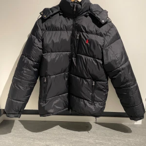 Svart dunjacka från Ralph Lauren - Snygg svart dunjacka från Ralph Lauren med dragkedja och ikonisk röd logga på bröstet. Jackan har en hög krage och praktiska fickor med dragkedja. Perfekt för kyliga dagar. Storleken är L och den sitter som L. Pris kan diskuteras 