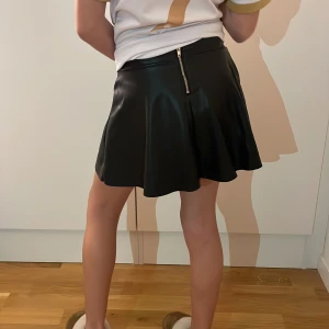 Svart kjol med dragkedja - Snygg svart kjol i skinn material. Storlek 116 men passar en som har storlek 146. Pris kan diskuteras!!