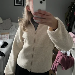 Beige teddyjacka med dragkedja - Mysig beige teddyjacka med dragkedja framtill. Perfekt för kyliga dagar med sin fluffiga och varma design. Jackan har en normal passform och långa ärmar.
