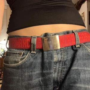 Rött/rosa bälte med metallspänne - Snyggt rosa bälte med ett brett metallspänne. Bältet har textdetaljer i en röd nyans som ger en cool look. Perfekt för att ge en färgklick till din outfit. Bältet är slitet med repor vilket ses i bilden men annars inga defekter.