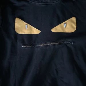 Svart hoodie från Fendi med ögonmotiv - Snygg svart hoodie från Fendi med ett unikt ögonmotiv på framsidan. Hoodien har en dragkedjedetalj som ger den ett coolt utseende. Perfekt för en stilren och avslappnad look. (Priset discutetas)