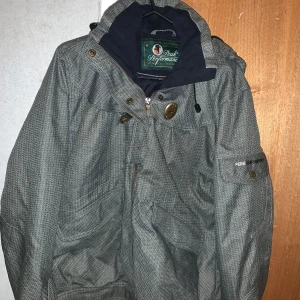 Grå jacka från Peak Performance - Snygg grå jacka från Peak Performance med en avtagbar huva och dragkedja. Jackan har knappar och en praktisk ficka på ärmen. Perfekt för kyligare dagar.