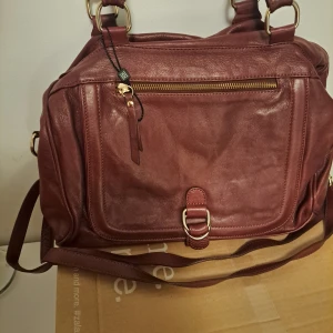 Vinröd axelväska med dragkedja - Snygg vinröd axelväska i skinn med praktisk dragkedja och justerbar axelrem. Väskan har flera fack och detaljer i guld, perfekt för att hålla ordning på dina saker.