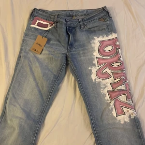 Blå jeans med graffiti-design från Replay - Coola blå jeans från Replay med unik graffiti-design i rosa och vitt på benen. Väldigt fina men färgen är smått sliten så därför ett billigt pris. Som sagt från replay och storlek 26-27. Jag köpte dem från Sellpy.