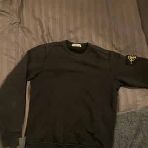 Svart sweatshirt från Stone Island - Säljer en svart sweatshirt från Stone Island med det ikoniska märket på ärmen. Tröjan har en klassisk rund hals och långa ärmar. Perfekt för en stilren och avslappnad look. Ända defekten är två hål på underärmen men inget som synd vid användning, skriv vid frågor och fler bilder 