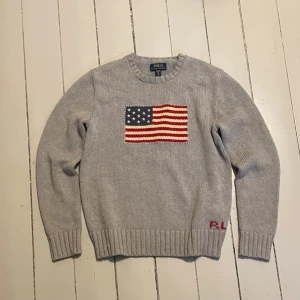 Grå stickad tröja från Polo Ralph Lauren - Säljer en stilren grå stickad tröja från Polo Ralph Lauren med ett amerikanskt flaggmotiv på bröstet. Tröjan har långa ärmar och ribbstickade muddar vid ärmslut och nederkant. Perfekt för kyliga dagar!