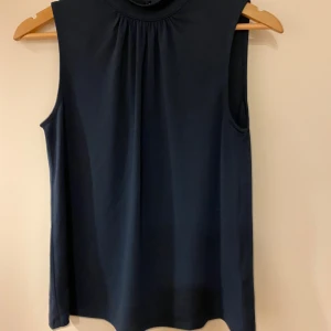 Mörkblå ärmlös topp - Snygg mörkblå ärmlös topp med hög hals och veckad detalj framtill. Perfekt för en stilren look.