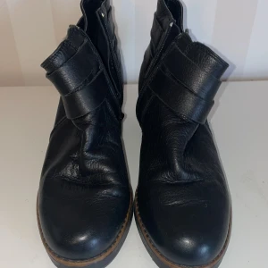 Nyskick Svarta boots med spännen - Snygga svarta boots i skinn med klack och dubbla spännen i nyskick endast testade 2-3 gånger . De har en dragkedja på sidan för enkel på- och avtagning. Perfekta för en stilren look.