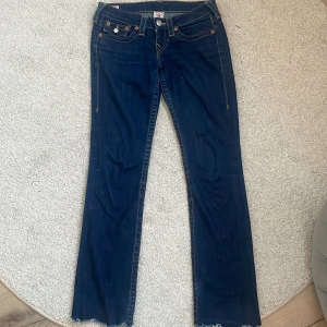 Mörkblå jeans från True Religion - Snygga mörkblå jeans från True Religion. De har klassiska femfickor och en unik broderad detalj på bakfickorna. Tillverkade i USA, dessa jeans är perfekta för en avslappnad look. Avklippta längst ned på benet!