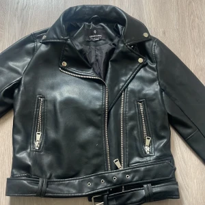 Svart skinnjacka med bälte - Säljer en snygg svart skinnjacka från stradivarius. Perfekt för höst och vår! Jackan är i nyskick och har långa ärmar.
