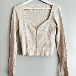 Beige ribbad tröja med knappar - Säljer en superfin beige ribbad kofta med knappar framtill. Den har en v-ringad design och långa ärmar, perfekt för både vardag och fest. Tröjan är croppad och ger en snygg och stilren look.