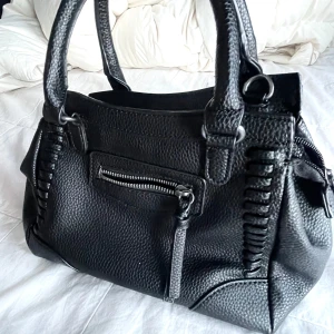 Svart handväska med dragkedja inkl band - Säljer en snygg svart handväska i skinn med coola detaljer. Väskan har en praktisk dragkedja och ett rymligt inre med flera fack. Perfekt för både vardag och fest!