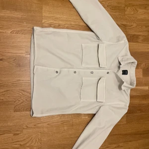 Vit overshirt från Zara - Säljer en stilren vit overshirt från Zara. Den har långa ärmar och två stora bröstfickor med lock. Skjortan är försedd med knappar framtill och har en klassisk krage. Perfekt för lager-på-lager-stil.