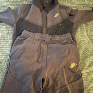 Nike tech tracksuite - Säljer ett par grå mjukisbyxor från Nike med elastisk midja och snörning. Byxorna har en gul Nike-logga på benet och är perfekta för en avslappnad stil. De är tillverkade i ett mjukt material för extra komfort.