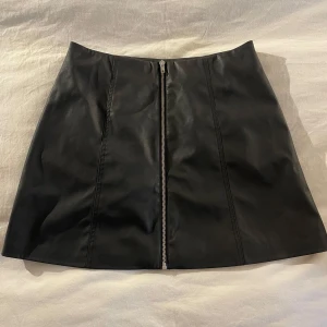 Svart kjol med dragkedja från H&M - Snygg svart kjol från H&M med en framträdande dragkedja framtill. Kjolen har en stilren design och är perfekt för en trendig look. Tillverkad i ett material som ger en skinnliknande känsla.