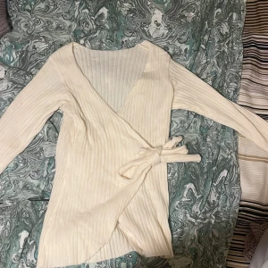 Vit ribbad omlottkofta - Säljer en stilren vit ribbad omlottkofta med knytband i midjan. Perfekt för en elegant och bekväm look. Koftan har långa ärmar och är gjord i ett mjukt material som passar bra till både vardag och speciella tillfällen.