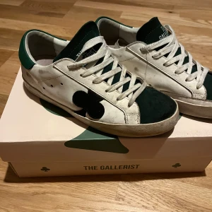 Vita och gröna sneakers från The Gallerist - Snygga vita sneakers med gröna detaljer från The Gallerist dom är använda men  är i bra skick , skorna kommer med låda dom är lite slitna vid sulan . Skorna har en svart klöverdetalj på sidan och vita snören. 