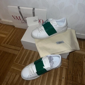 Valentino Garavani  - Snygga vita sneakers från Valentino Garavani med ett iögonfallande grönt band över snörningen. Skorna har en klassisk design med rund tå och är tillverkade i skinn. Perfekta för att ge en stilren touch till din outfit. Fått nyligen men passar tvyär inte. Ny pris 7.899, PRIS KAN DISKUTERAS VID SNABB ÄFFER