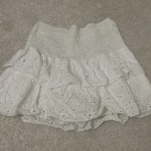 Vit kjol med volanger - Charmig vit kjol med volanger och broderade detaljer. Perfekt för en somrig look med sin luftiga design och elastiska midja. Kjolen har en kort längd och ger en romantisk känsla. Köpt i Spanien 