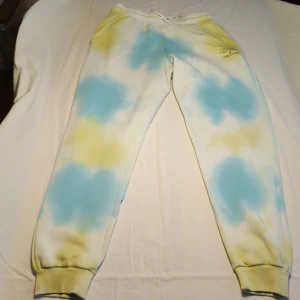 Färgglada mjukisbyxor från Continu - Säljer ett par mjukisbyxor från Continu i en cool tie-dye stil med blå och gula nyanser. Byxorna har en elastisk midja med snörning och ribbade muddar vid bensluten. Perfekta för en avslappnad look!