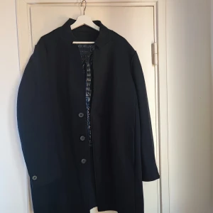 Svart kappa med inbyggd fodrad väst - Elegant svart kappa med inbyggd fodrad väst och knappar framtill. Den har en klassisk design med långa ärmar och en stilren look. Perfekt för kyligare dagar och ger en sofistikerad touch till din outfit.