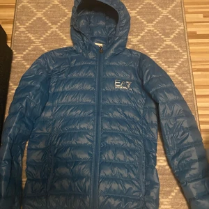 Blå dunjacka från Emporio Armani - Säljer en snygg blå dunjacka från Emporio Armani med huva och dragkedja. Jackan har en quiltad design och EA7-logga på bröstet. Perfekt för kyligare dagar!