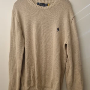Beige stickad tröja från Polo Ralph Lauren - Beige stickad tröja från Polo Ralph Lauren med det klassiska logotypbroderiet på bröstet. Tröjan har en rund halsringning och ribbade muddar vid ärmslut och nederkant.